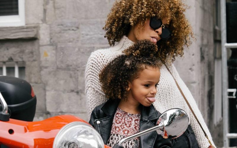 mother and daughter stick their tongues out while looking at themselves in a motorcycle mirror parked in front of a building